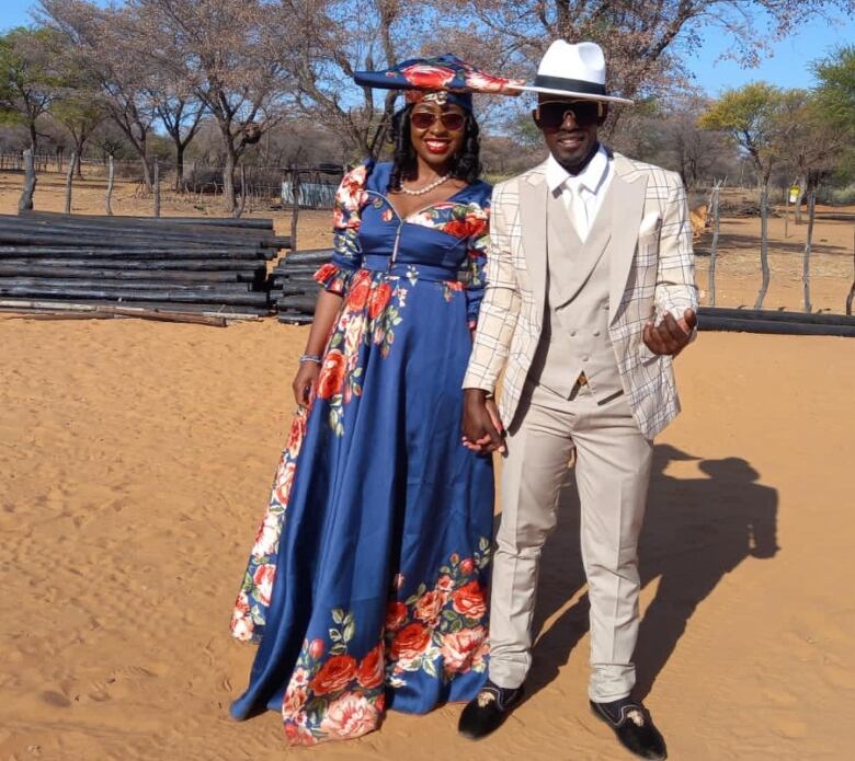 A man wearing a tan-coloured three-piece suit holds hands with a woman wearing a flowing blue gown patterned with red flowers. 