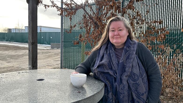 A woman sits at a table outside, smiling at the camera while holding a cup of coffee. In the background is a green gate and a parking lot.