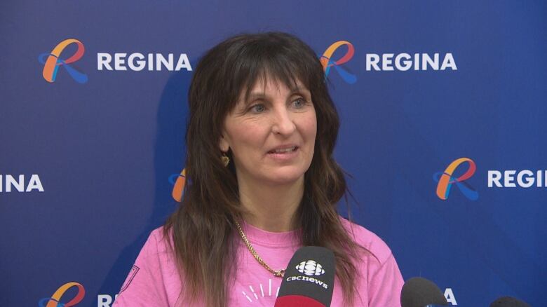 An older woman wearing a pink t-shirt and gold necklace with long, dark brown hair, stands behind two microphones, in front of a City of Regina backdrop, to address reporters.