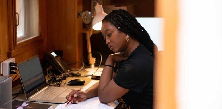 A woman sitting at a desk is looking at printed notes and a laptop.