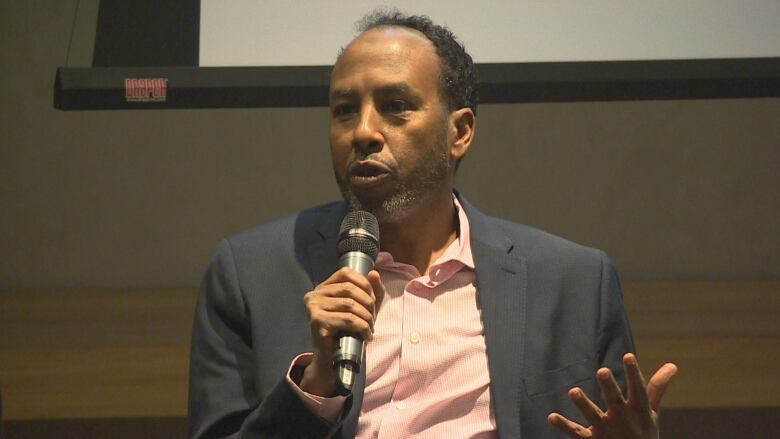 A man speaks into a microphone he holds while sitting down.