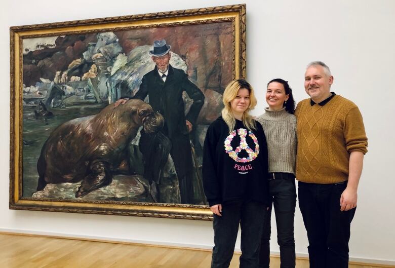 Three people stand in a museum in front of a painting.