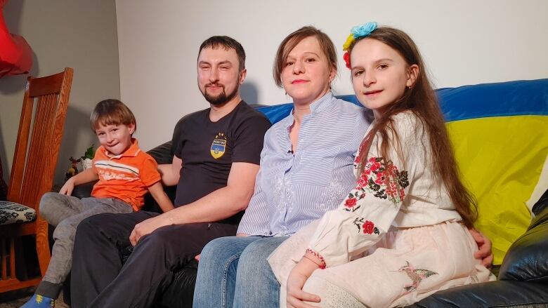 A family of four sits on a black leather couch. The yellow-and-blue Ukrainian flag is draped over the back of the couch. A little boy sits on his fathers lap. The daughter sits with proper posture next to her mother. There is a wooden chair in the background.