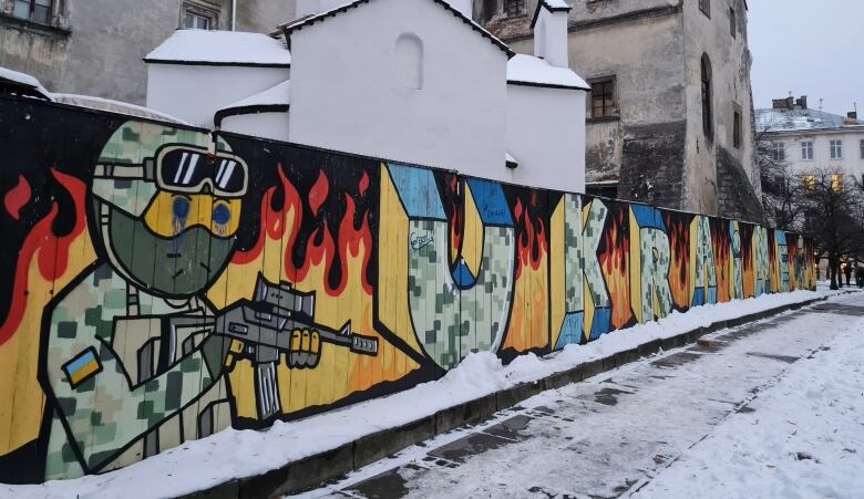 A graffitied mural shows a cartoon Ukrainian soldier carrying a rifle. The word 'Ukraine' is spelled out, with each letter being painted in army camouflage. There are flames painted in the background.