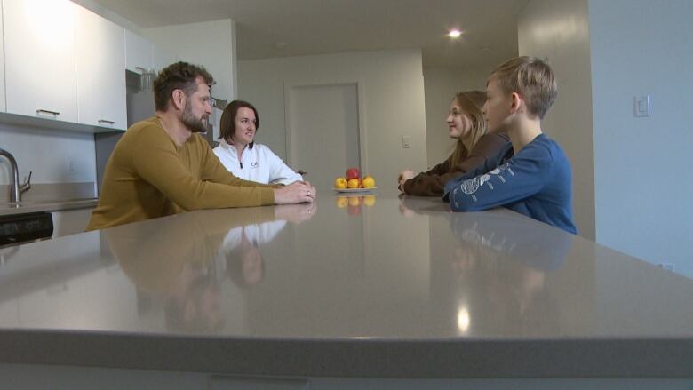 A family of four sits across from each other at a kitchen island. The mother and father on one side, the brother and sister on the other. A bowl of fruit sits on the counter between them. They are in an apartment with white walls and doors, but the counter is grey.