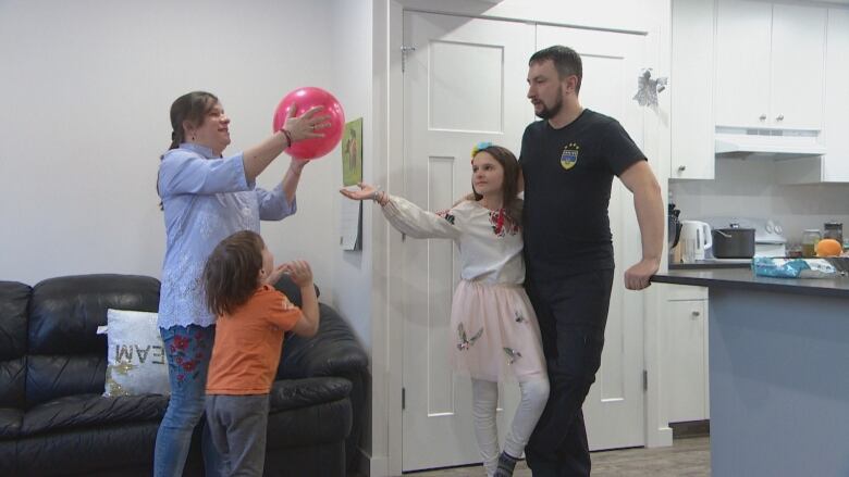 A family of four is standing in their living room. The mother is holding up a red balloon. Her son, standing beside her, is looking up toward the balloon. Her daughter is standing across from her; the girl's left arm is wrapped around her father and her right arm is reaching toward the balloon. The father watches, holding his daughter.