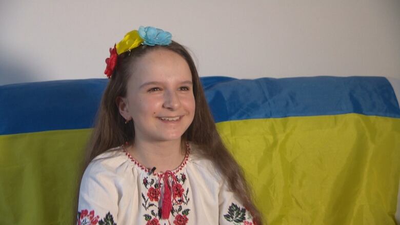 A young girl is smiling while sitting on a couch. The Ukraine flag is draped over the back of the couch, behind her. She is wearing a white dress, with an embroidered pattern of roses near the neck. She is wearing a clip in her hair with fake flowers of blue, yellow and red.