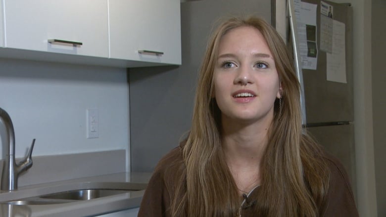 A teenage girl with straight blonde hair, wearing a brown zip-up sweater, is sitting in a kitchen and speaking.