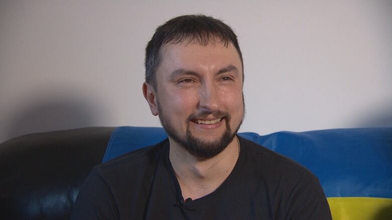 A man with dark hair and a dark beard, wearing a black shirt, is smiling. He sitting down on a couch.