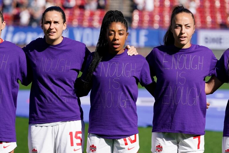 Three female soccer players stand with arms linked, wearing shirts that say 