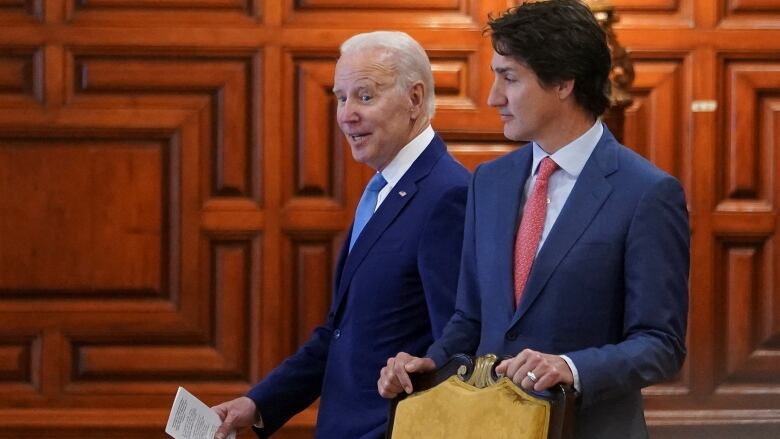 Two men in suits walk together. The older one, with white hair and a blue tie (Joe Biden) looks mid-sentence. The younger one, with touselled brown hair and a red tie (Justin Trudeau) raises his eyebrows.