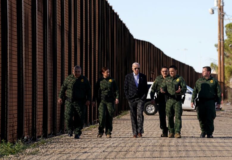 A man with white hair and aviator sunglasses wearing a dark suit, collar button open (Joe Biden) walks with what appear to be soldiers in green uniforms near a tall fence.