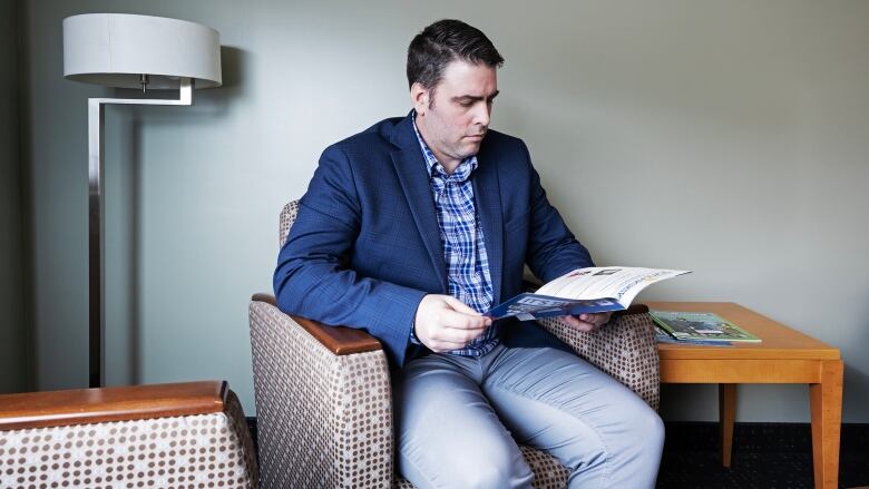 A man in a blue blazer sits in a chair, reading a pamphlet.