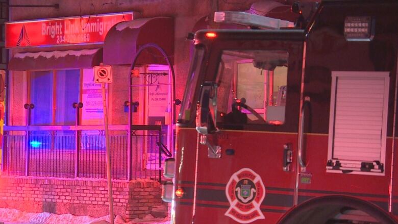 The front end of a fire truck is seen at right in a photo coloured in red from the emergency lights. A business is seen at left of the photo with a metal railing around a patio at the entrance.