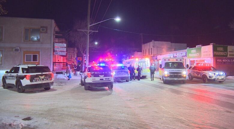 Police cars, ambulances and fire trucks are parked on a snowy street at night, their coloured lights flashing off buildings.