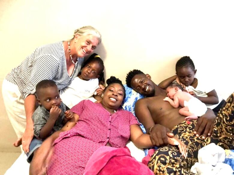 A smiling woman wearing pink pajamas lies in bed next to a man with a newborn baby on his bare chest. Three other children and a grey-haired lady are also smiling for the camera.