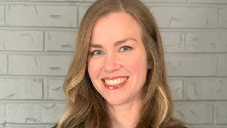 A woman with long light brown hair and blue eyes stands in front of a light coloured brick wall with her arms crossed. She is wearing a long sleeve black shirt.