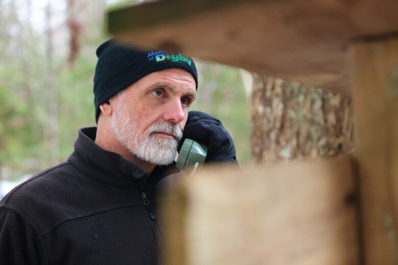 A man wearing a tuque is outside in a snowy forest. He is holding a rotary phone receiver to his ear.