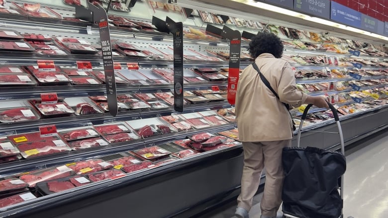 A customer in a brown jacket looks at meat in a grocery store aisle.