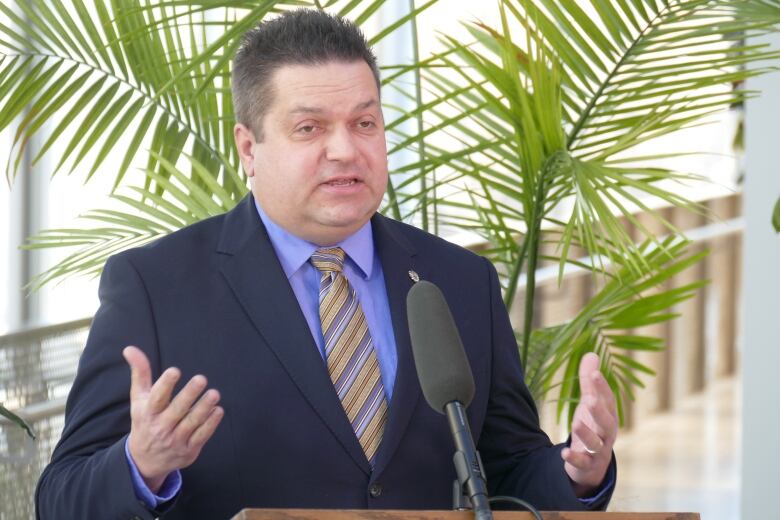 Man stands at podium with arms raised to chest and palms facing up.