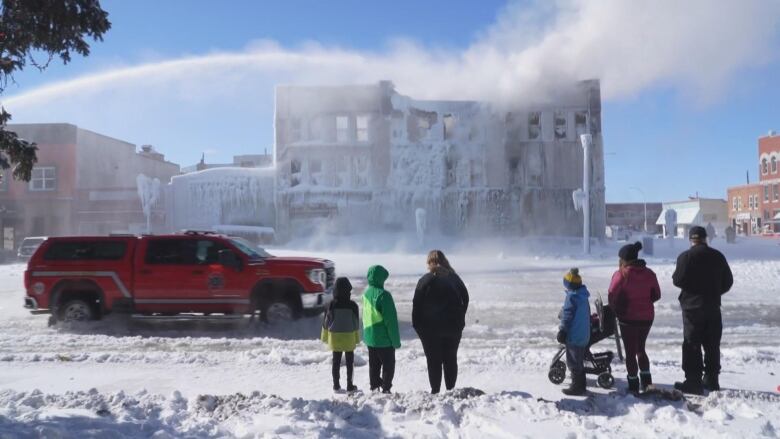 a red truck is parked in front of a frozen three floor building. a person off camera is spraying it with a hose. six people stand on the sidewalk across from it watching