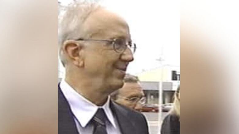 A man with white hair in a black suit and glasses is pictured.