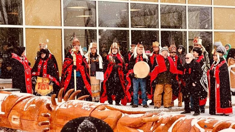 A group of people in Indigenous clothing are holding drums in front of a building and behind a laid-down totem pole.