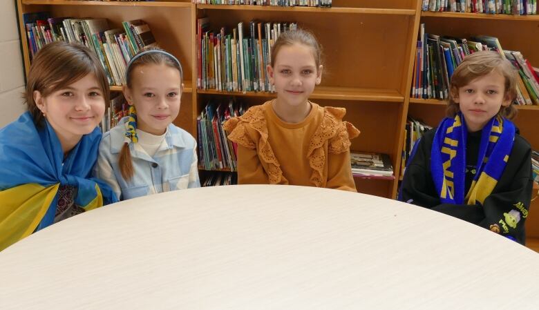 Four children, one of whom wears a Ukrainian flag draped over her shoulders, sit at a round table in a classroom, smiling for the camera. 