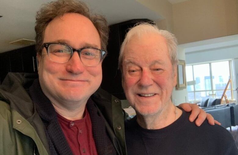 Two men stand looking into the camera, one with his arm around the other.