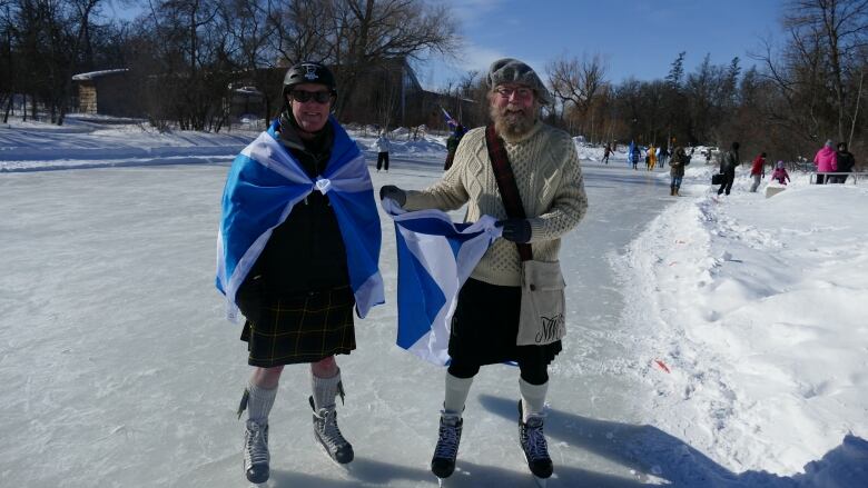 Two men in kilts and ice skates stand on the ice and smile to the camera.