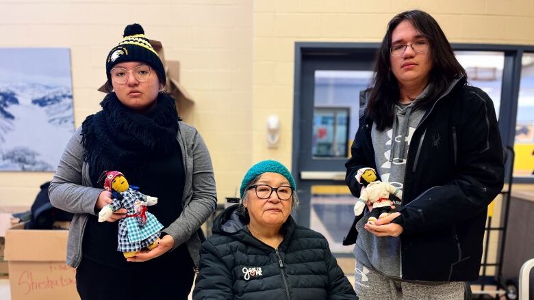 A senior woman sits in a chair while two teenagers stand on either side of her. The two teenagers are each holding a traditional doll. 