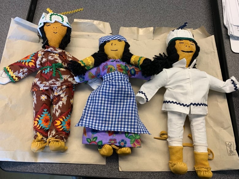 Three dolls made out of faux caribou skin are shown on a table. One has a patterned red outfit, one has a blue plaid dress, and the other is in traditional white clothing. 