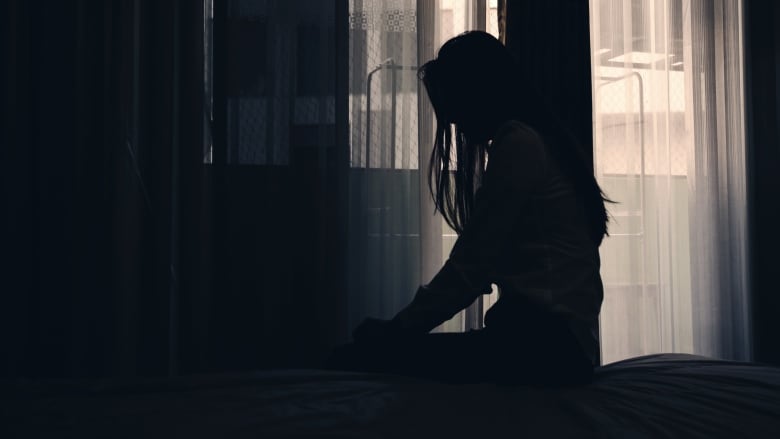 A woman sits on a bed, silhouetted by light coming from an open window.