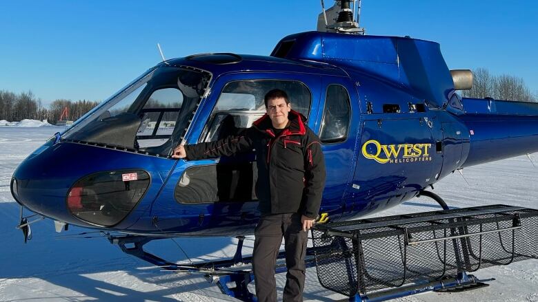 Man in a black and red winter coat stand in front of a blue helicopter in a snowy field. 