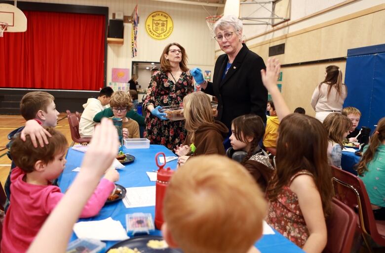 A woman with short grey hair and glasses wears blue plastic gloves and a dark blazer as she talks to a group of children seated at a table in a school gymnasium. Two of the children have their hands raised as if to ask a question.