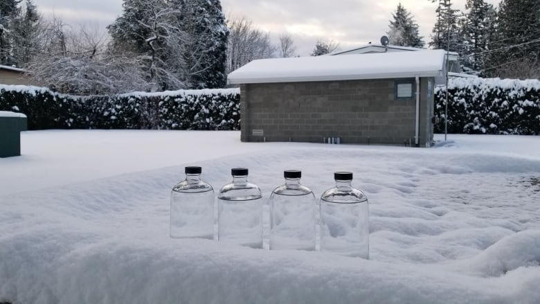 Four glass jugs filled with water sit foreground in snow, with a brick building in the rear of the photo.