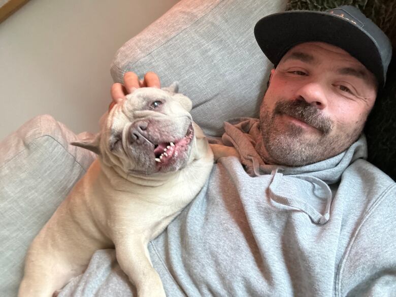 A man in a baseball hat lies on a couch while patting a French bulldog on the head.