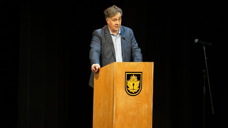 A middle-aged man in a blue jacket stands behind a podium.