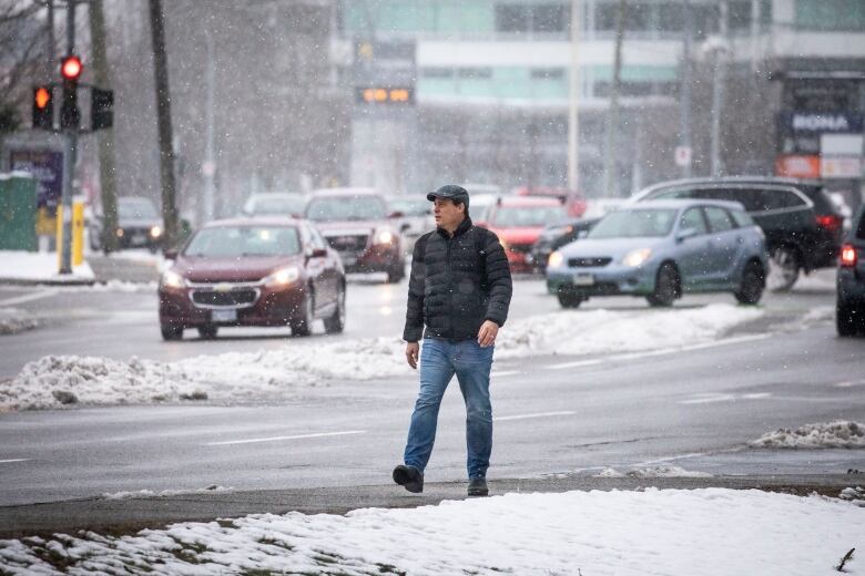 A man in a ball cap and black puffer jacket walks down a snowy sidewalk. Traffic is on the road in the background.