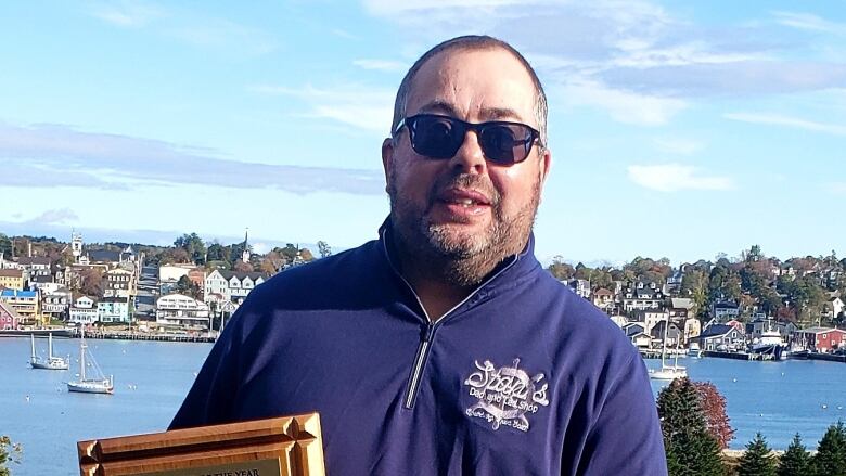 A man with sunglasses, beard and a blue sweater stands atop a hill with Lunenburg harbour and the colourful waterfront spread out behind him.