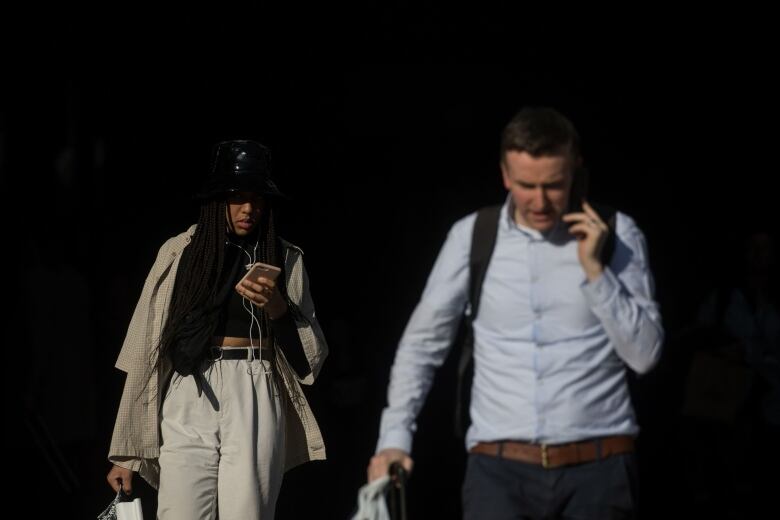 A woman in the background and a man in the foreground are shown using their cellphones while walking outside.