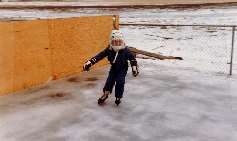 A boy wearing a hockey helmet, hockey gloves, a snowsuit and ice skates on an outdoor rink. 