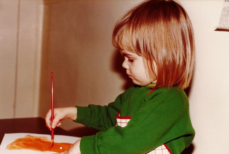 A toddler focuses intently while applying bright orange paint to a piece of paper.   