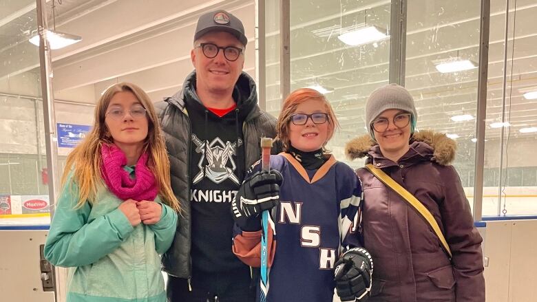 Alt text: A woman wearing a brown winter jacket with a fur hood smiles while standing next to a man, a girl and a boy in a navy blue hockey jersey and holding a hockey stick. 