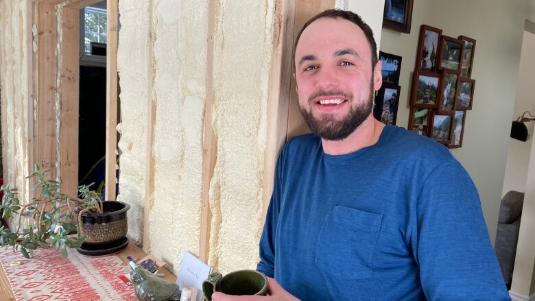 A man holding a coffee cup standing in front of an exposed wall with spray foam in it.