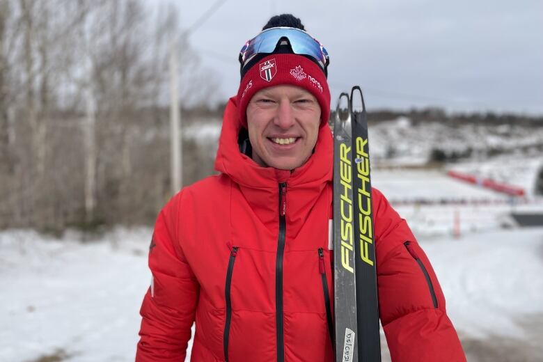 Man in red ski jacket holds up his skis with snow in the background.