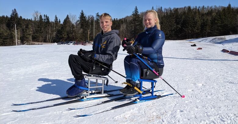 A young man and woman in ski clothes sit on metal sit-skis during a practice session.