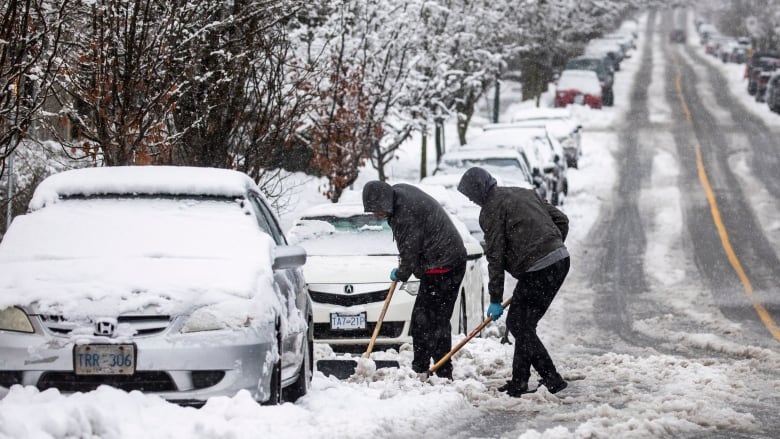 Two people with snow shovels dig out a silver car parked on a residential street.