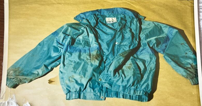 A teal windbreaker-type jacket, with some dark stains on it, is spread out on a wooden table