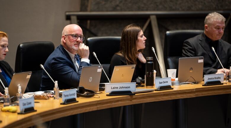 Four people sit at a council table, with one holding his chin in his hands and looking off into the distance.